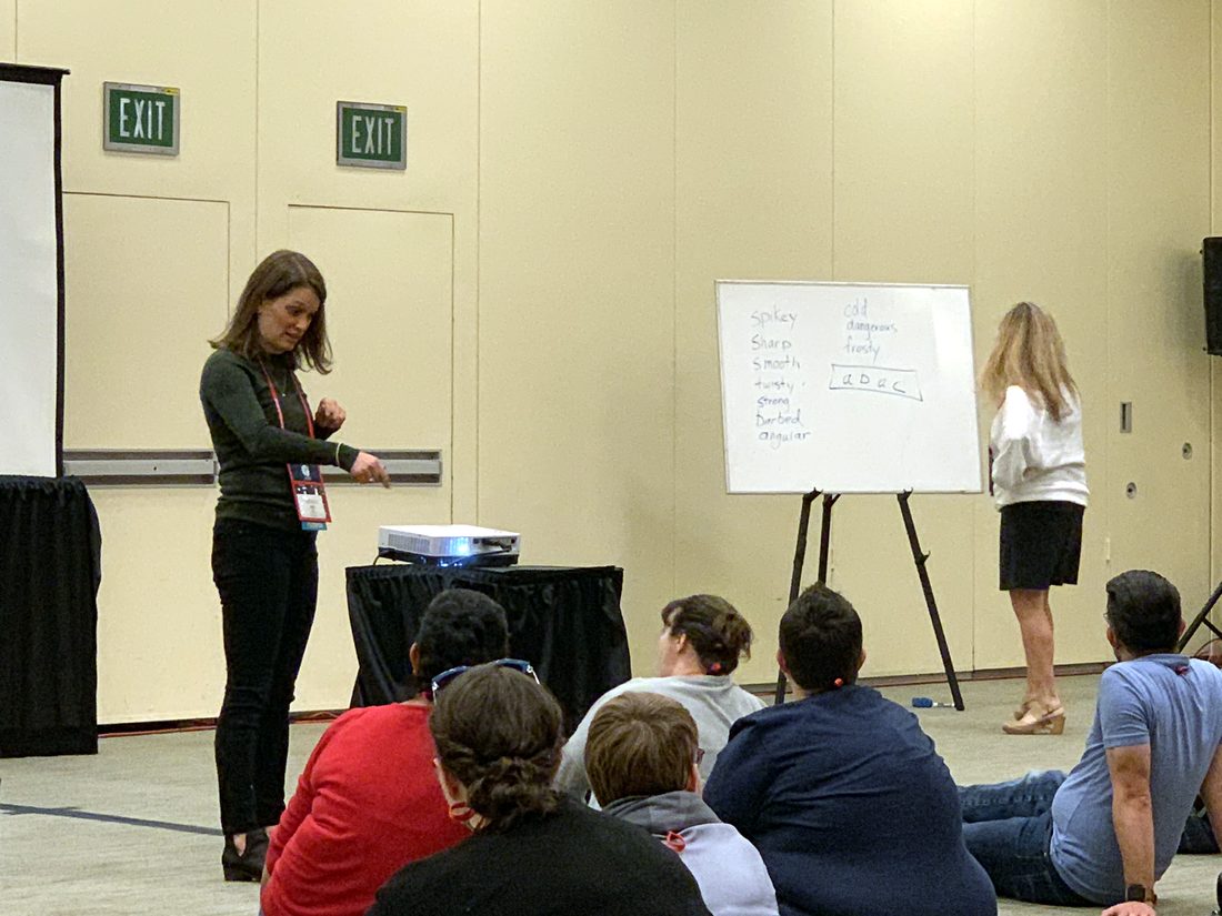 Beth Brown and Diana Hawley teaching a session for music educators, a whiteboard of descriptive words between them, teachers seated on the floor